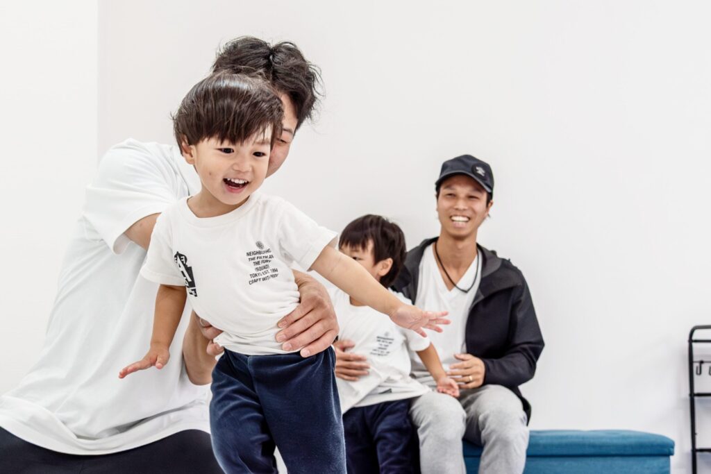 笑顔で体を動かす子どもと、それを優しく支える講師、見守る保護者の様子